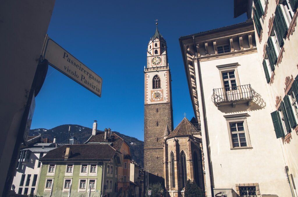 La torre campanaria del Duomo di Merano con orologio, sullo sfondo le montagne dell’Alto Adige.